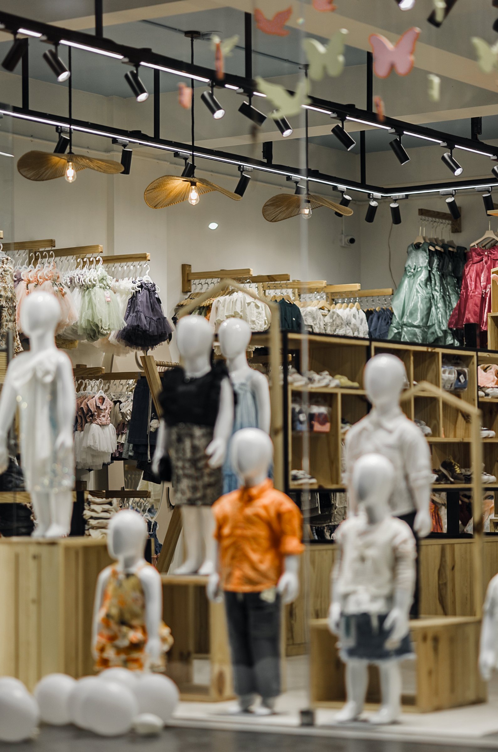 Portrait view across child mannequins toward pale wood racks and a plant-lined window