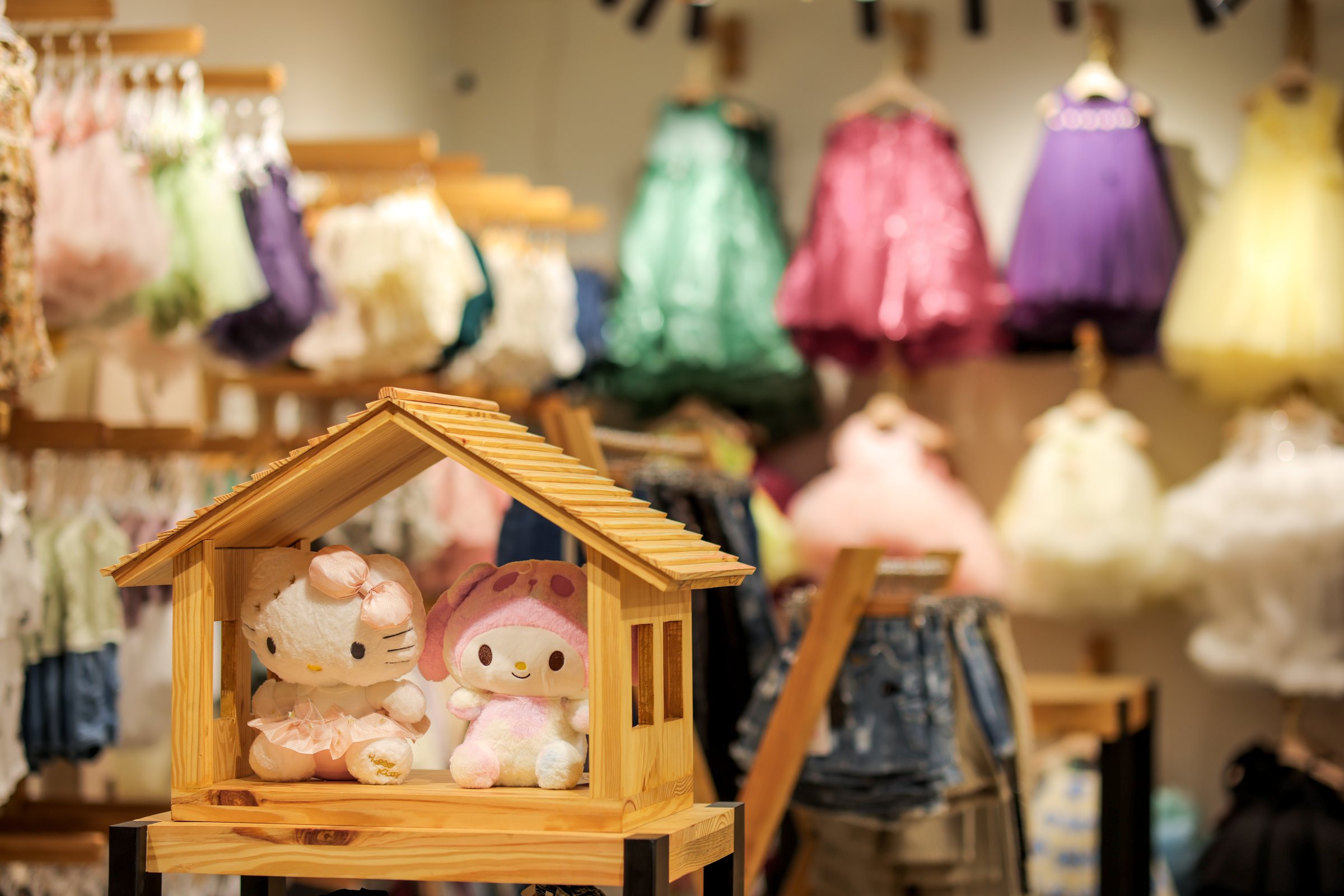 Two plush characters seated in a miniature wood house with colorful dresses behind