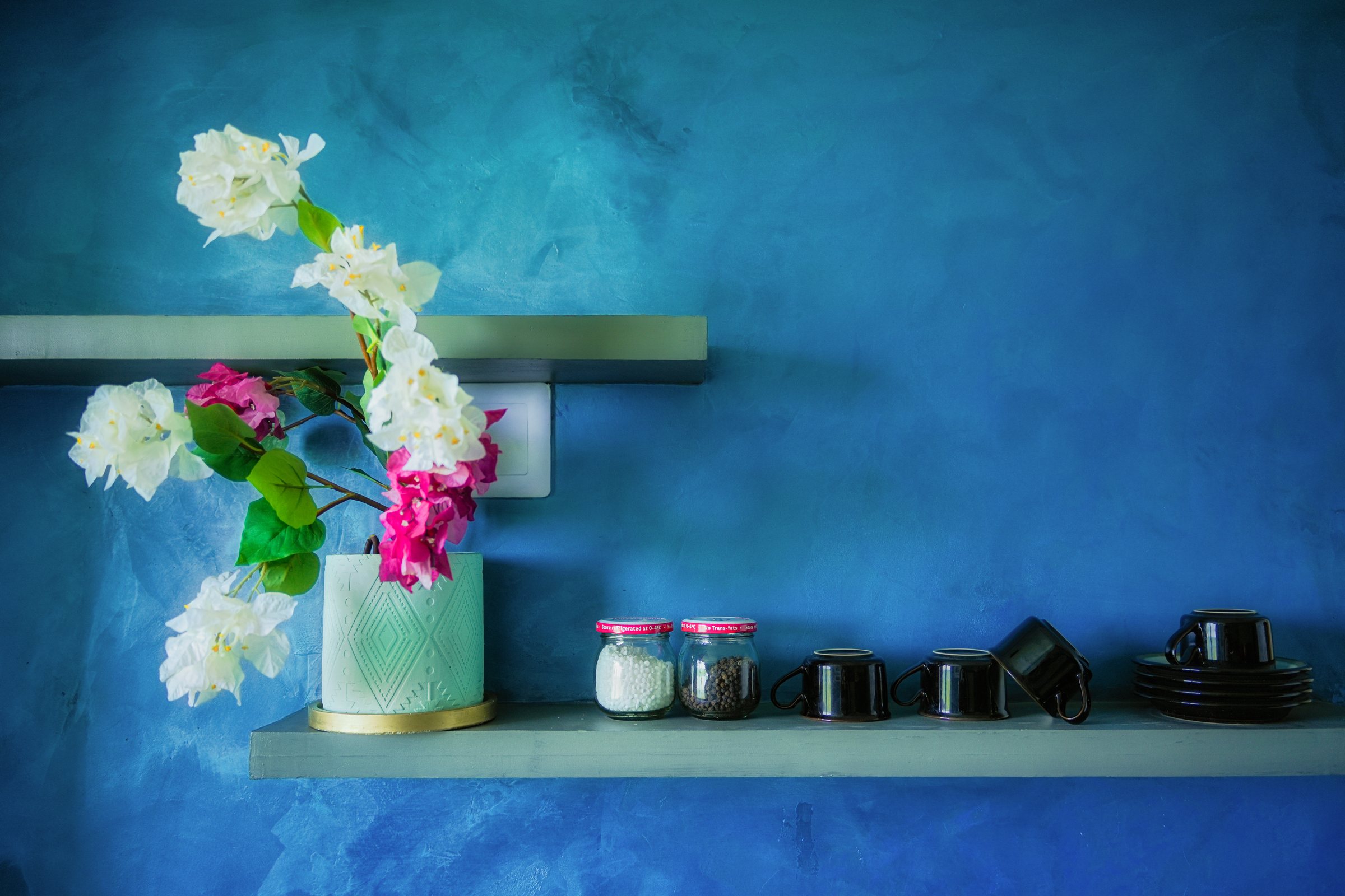 Black cups, jars and flowers arranged on a floating shelf against a textured blue wall